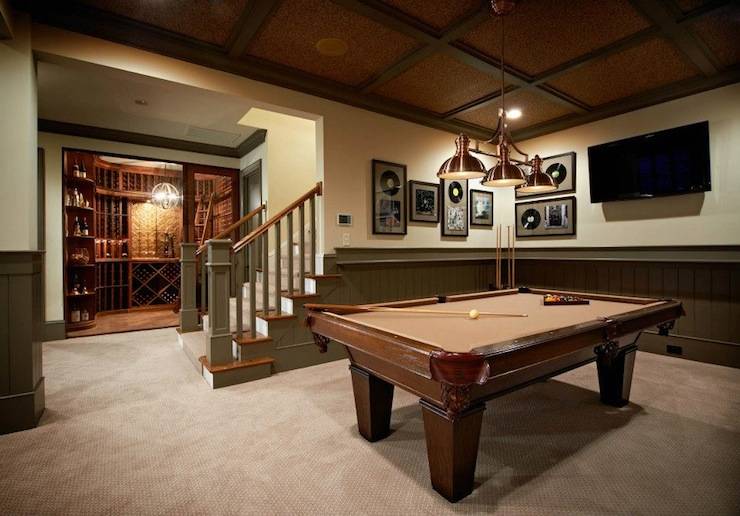 Fabulous basement games room with separate wine cellar. The basement features tongue and groove paneling in a khaki green with light yellow upper walls. The ceiling is coffered with dark stained beams and a dark brown center panel. Framed records and a flat screen tv are mounted on the walls behind the pool table. The traditional pool table is lit by a three light copper pendant. The wine cellar features hardwood floors and custom wine storage. The wine cellar is lit by an armillary style pendant.
