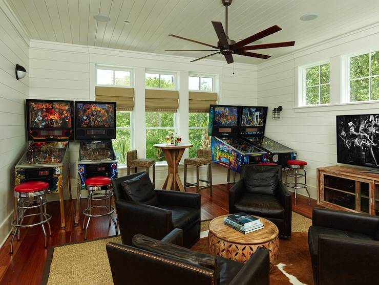 Dream Man Cave style games room with pinball machines and fifties style chrome bar stools flanking three windows. Below the windows stands a pub height wooden table paired with seagrass bar stools. In the foreground four espresso leather club chairs with nailhead trim surround a round wooden drum coffee table. The seating arrangement is anchored by a brown and white cowhide rug layered on top of a bound sisal rug over hardwood floors. To the right of the seating arrangement stands a distressed wood media console with flat screen tv. The room features white shiplap paneled walls and wood paneled ceilings. A contemporary ceiling fan adorns the ceiling over the space.