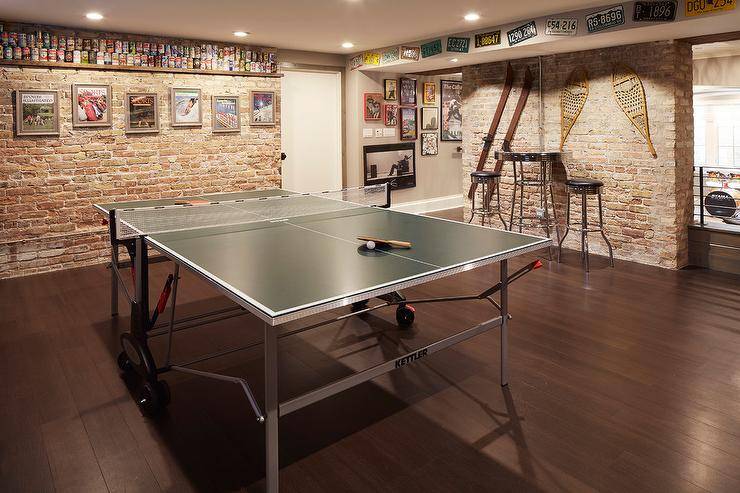 Basement man cave features a ping pong table placed in the center of a room clad in exposed brick and fitted with a long floating shelf showcasing stacked beer cans and fixed above framed photographs. A tall round pub tables seats two backless stools against a wall accented with antique sports gear and beneath a row of license plates.