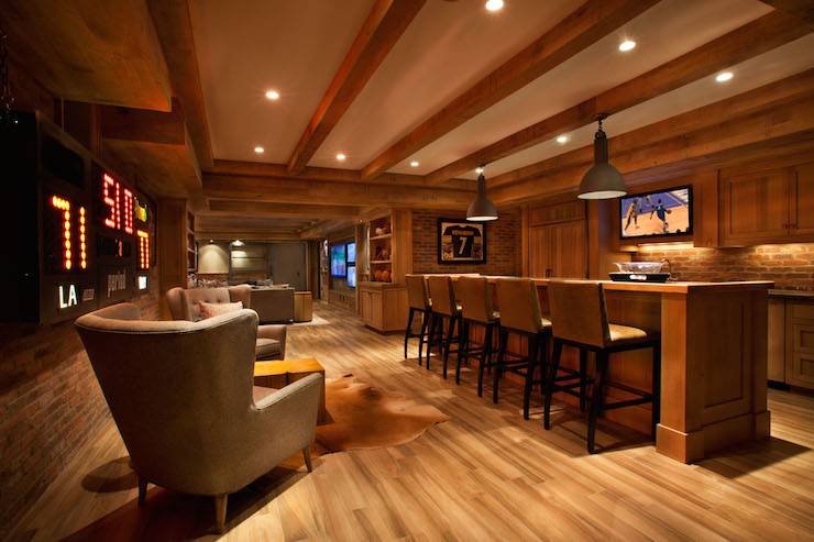 Basement man cave features wood ceiling beams interspersed with pot lighting and gray industrial pendants illuminating a bar island lined with leather barstools across from rustic wood paneled refrigerator next to flatscreen TV over wet bar sink framed by stained cabinets topped with dark countertops. Man cave in basement boasts exposed brick walls framing digital basketball scoreboard placed behind a pair of gray tufted wingback chairs atop wood floors.