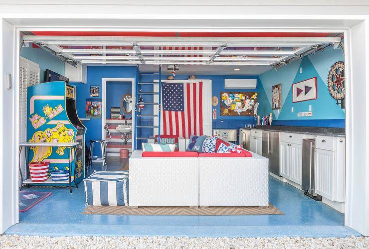 Converted garage man cave features a vintage arcade Ms. Pac-Man game in front of a white wicker sectional with red seat cushions and a matching coffee table. Blue walls, beadboard cabinets, and gray countertops include a kitchenette in the converted man cave space.