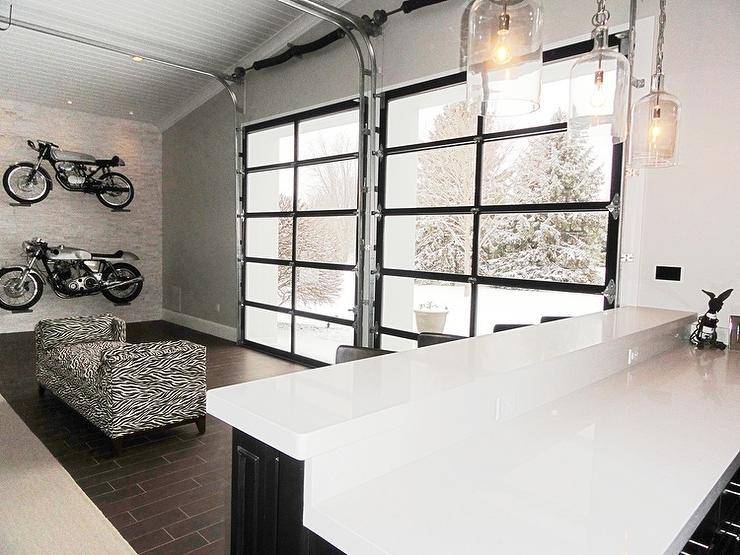 Man cave Boasts a vaulted ceiling over a stone clad walls lined with stacked display motorcycles surrounding a zebra bench facing glass garage style doors. A man cave bar features black cabinets topped with white quartz illuminated by Corsica 1 Light Pendants.