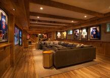 Basement man cave features wood ceiling beams interspersed with pot lighting over top half of walls painted beige and bottom half of walls clad in rustic paneling highlighting framed basketball jerseys illuminated by iron and glass globe light pendants. A U shaped gray sectional sofa is paired with side-by-side brown leather ottoman coffee tables as well as hex accent tables atop sand colored rug facing a flatscreen TV.