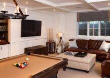 Fantastic basement features a coffered ceiling accented with a leather and iron chandelier illuminating a pool table facing a built-in bar nook. Basement ma n cave boasts a brown leather sofa and chairs facing a beige tufted storage ottoman facing a vintage trunk under a flatscreen TV.