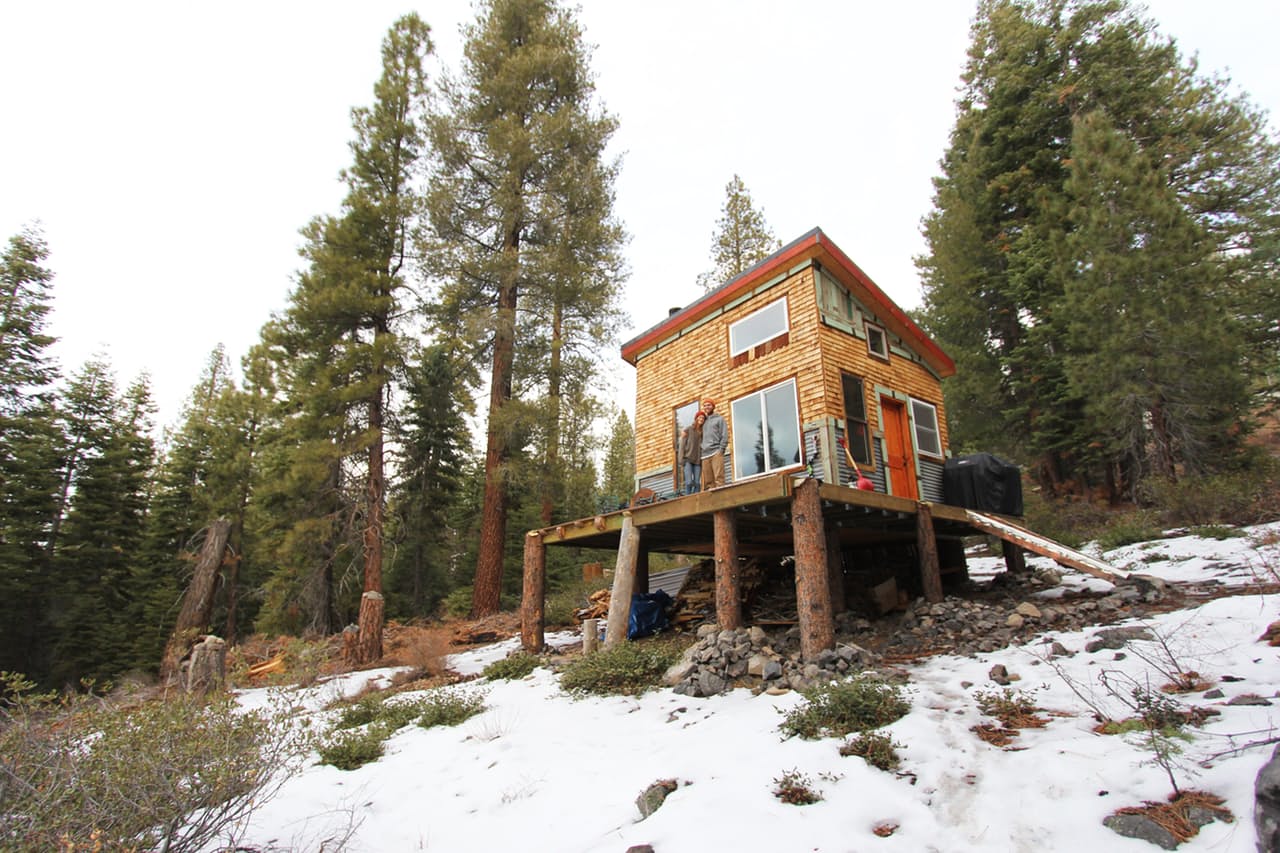 Couple posing in front a tiny home elevated on a slight hill covered in fresh snow.