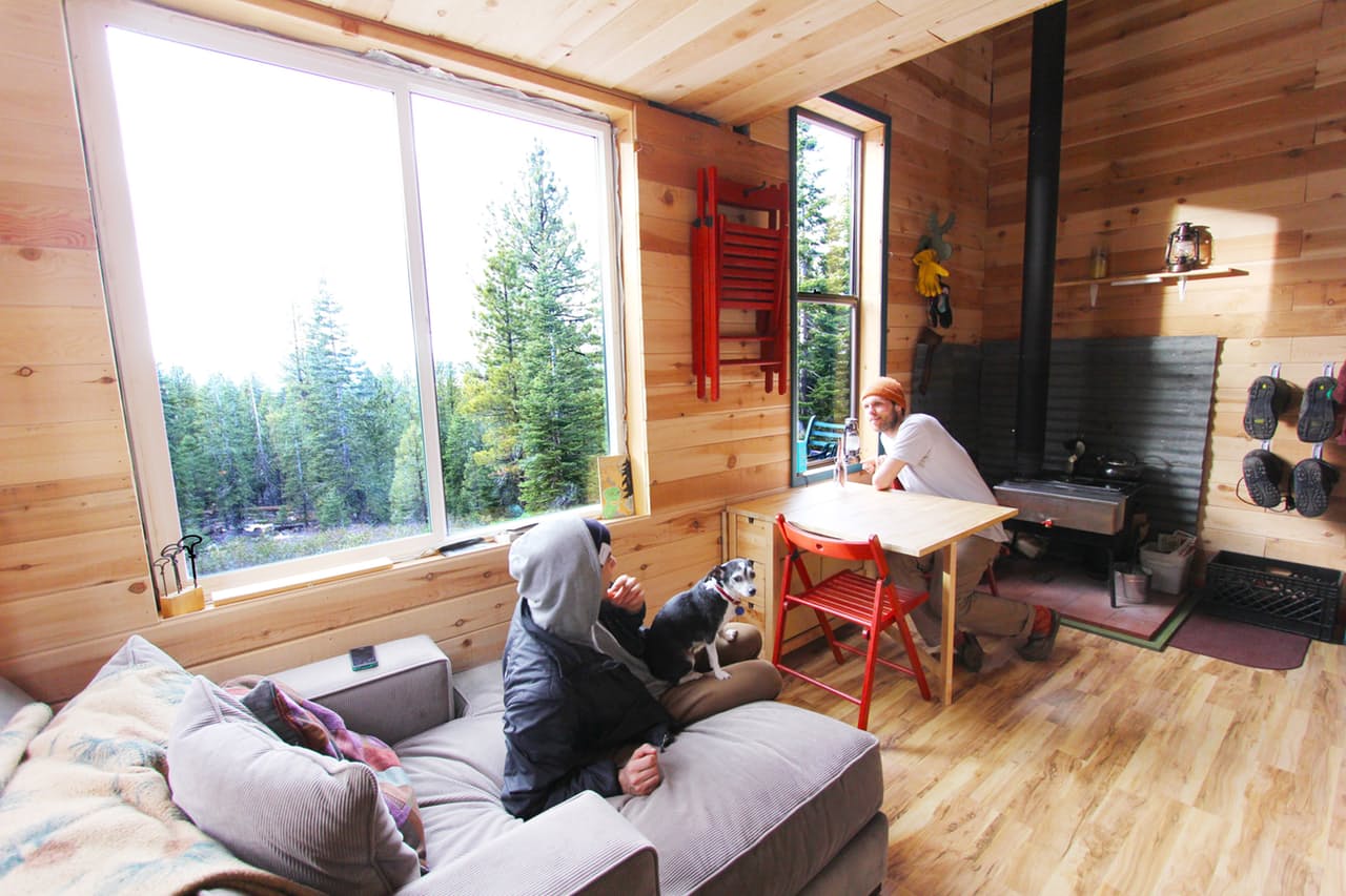 Two people are hanging out in a tiny home, one sitting on the couch with a dog while the other is leaning on a wood table near the window.