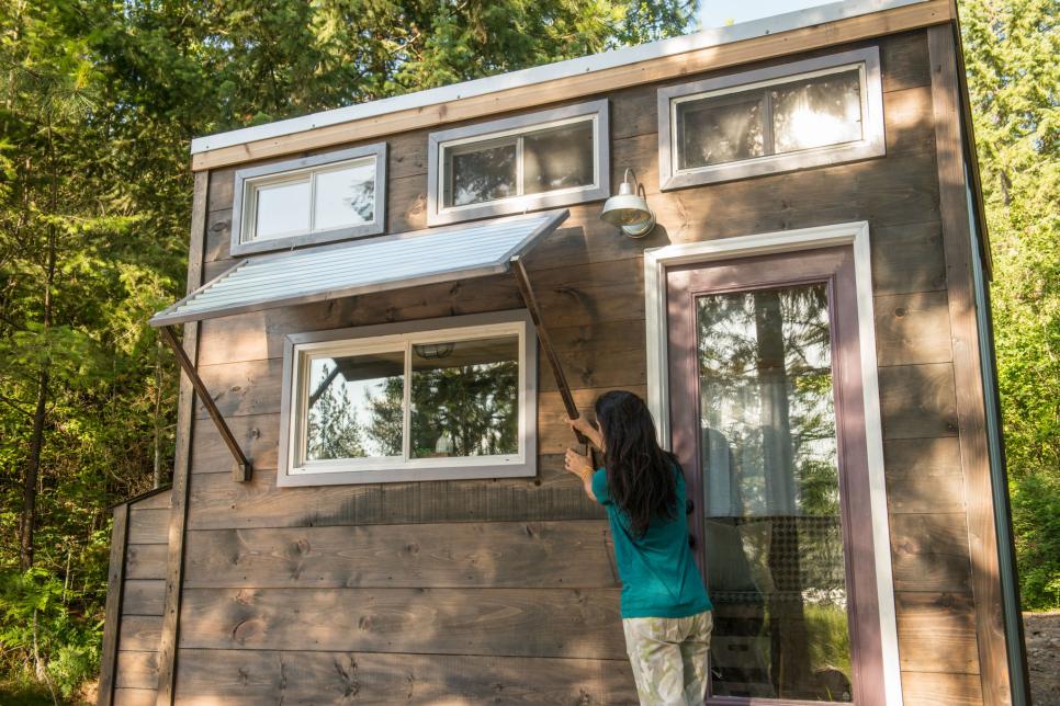 Person adjusting window shade on the exterior of a tiny home that has multiple windows and wood exterior finish.