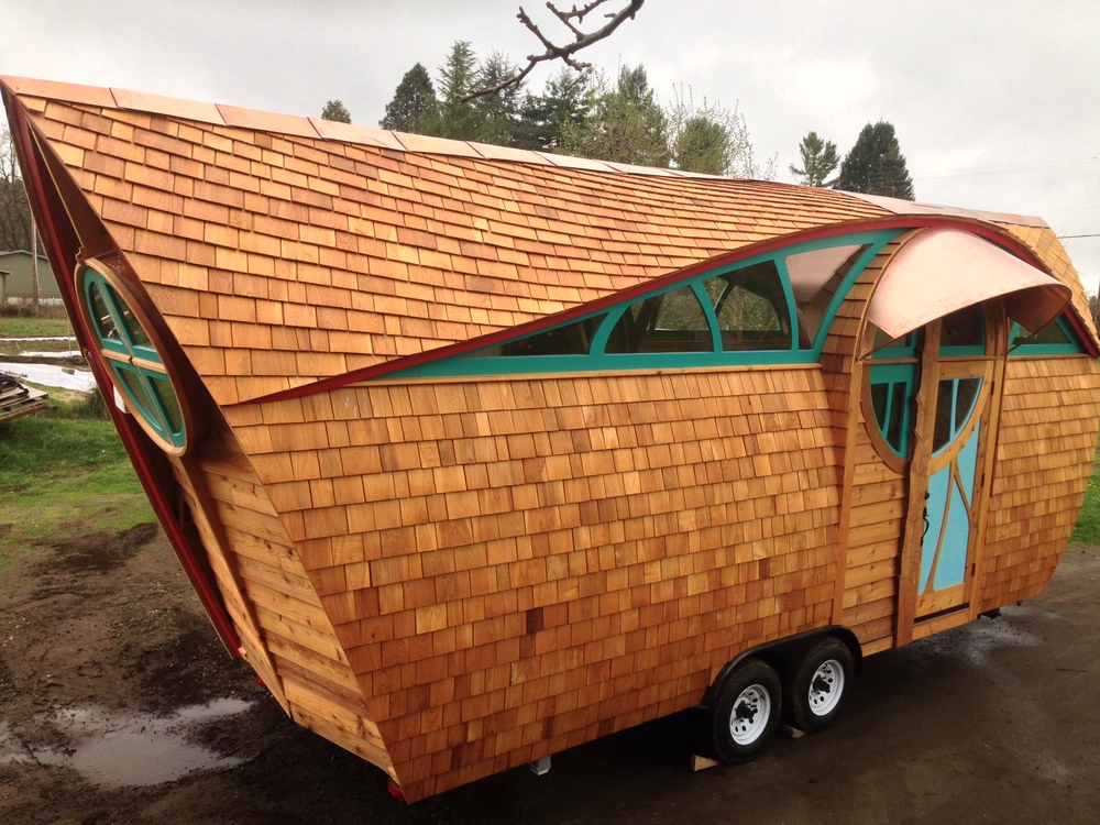 Uniquely shaped tiny home on a trailer frame, which has a roof made of shingles that spans almost the entire way down and has green accents.