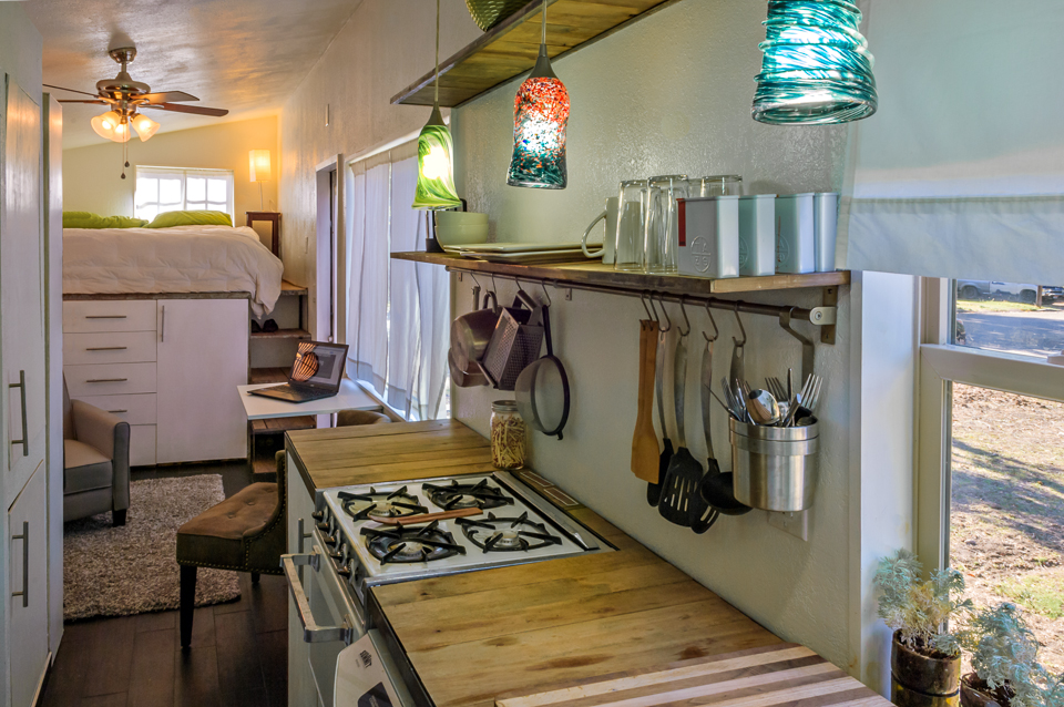 Interior of tiny home showcasing wood kitchen counter, gas stove, and white bed.