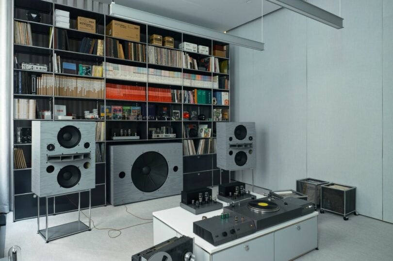 OJAS Listening Room at USM Modular Furniture with large modular bookshelf system filled with books, vinyl records, audio components and other miscellaneous items, with turntable and two tube amps set across a rolling platform storage. Room is decorated in all neutral gray decor.