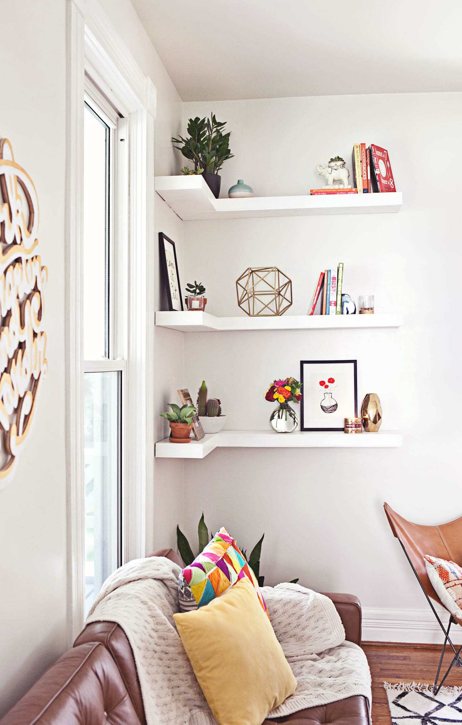 Three white corner-mounted floating shelves hold a small plant, pictures, and a uniquely shaped decor piece.