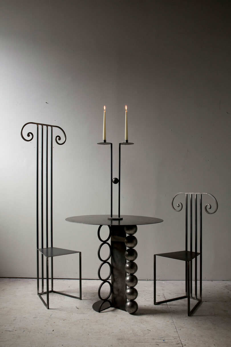 Two minimal metal chairs with seat backs that look like fluted columns staged with a table and candelabra.