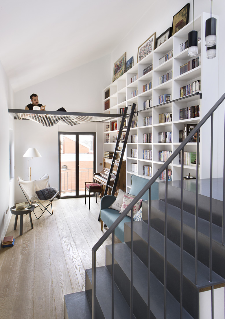 Man seems relaxed, engrossed in a book on a hammock inside a room with stairs and a large book shelf.