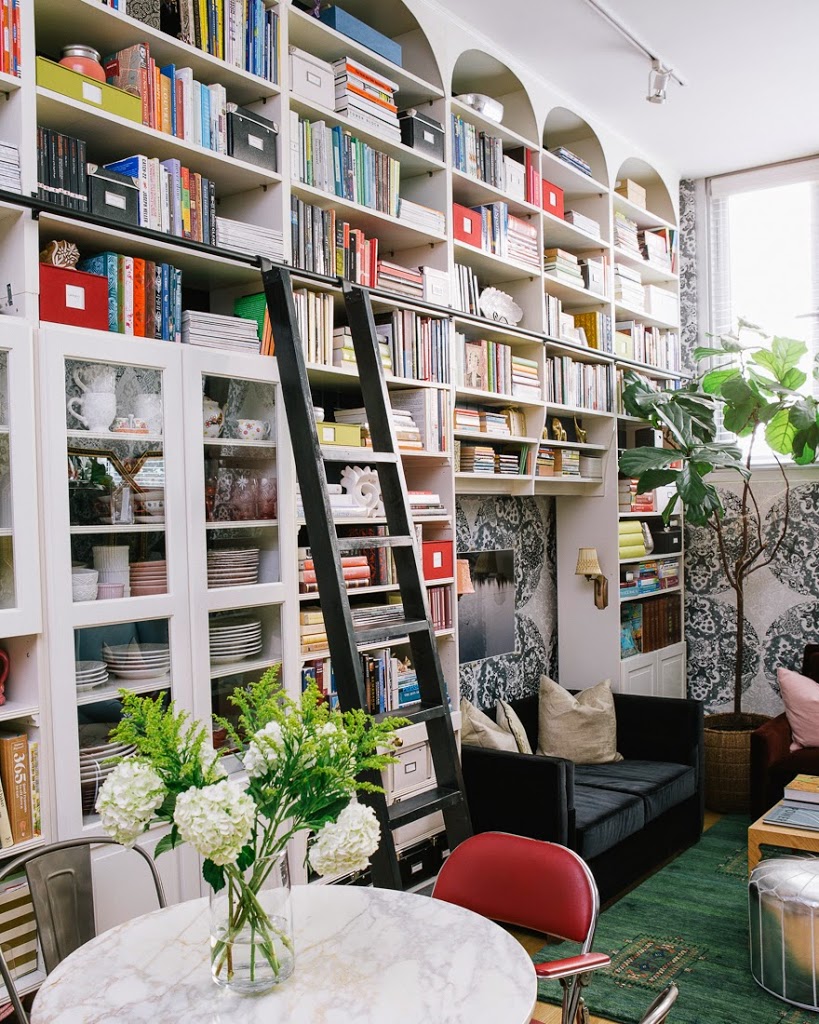 A cozy living room with a table adorned with a vase of flowers, surrounded by a red chair and a large book case.