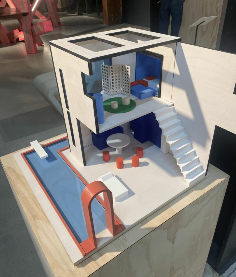 A modernist cube style dolls house on a plinth features two floors, an external staircase and a pool wrapped around two sides. 