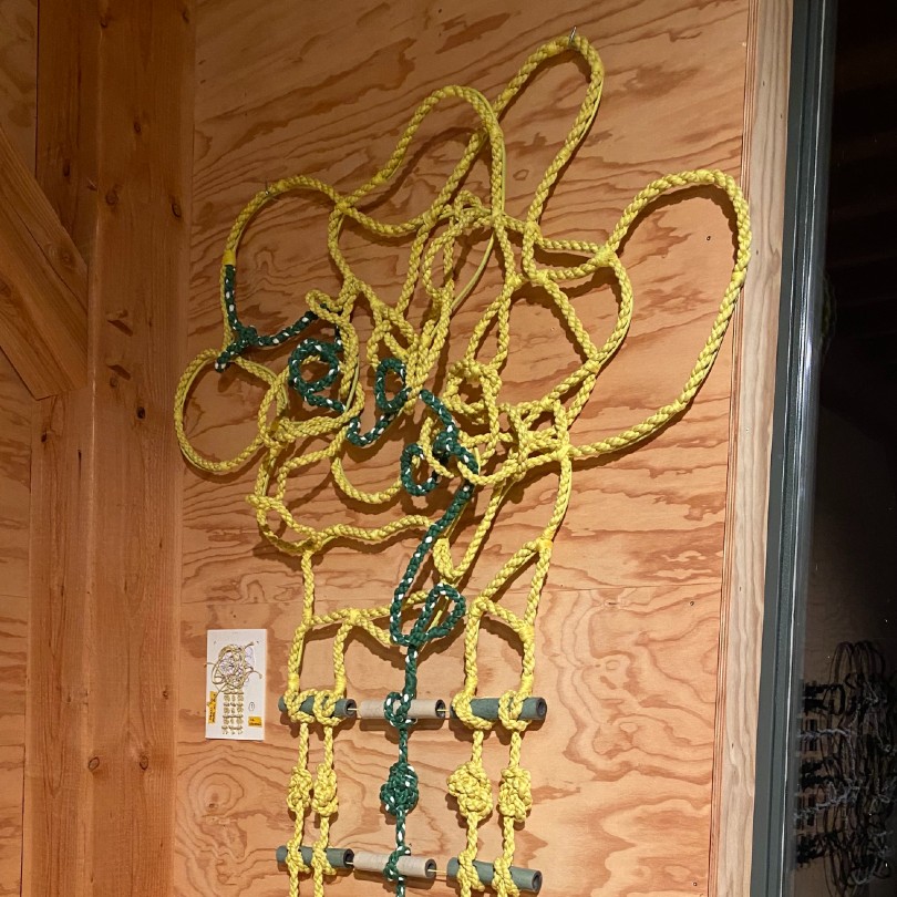 A knotted rope with horizontal ceramic bars starts in a chaotic form at the top and gradually resolves itself into a grid as it moved down. The whole thing is hung on a wooden wall next to a darkened window. 