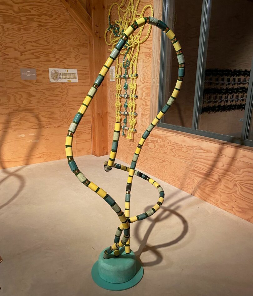 A sculptural form comprises a green hat-shaped base with a curvilinear loop and line emerging out of it that is made from a series of overzied tubular beads in various shades of yellow and green. 