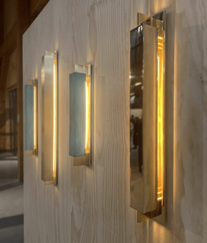 Four linear vertical brass light fittings are affixed to a wooden wall. 