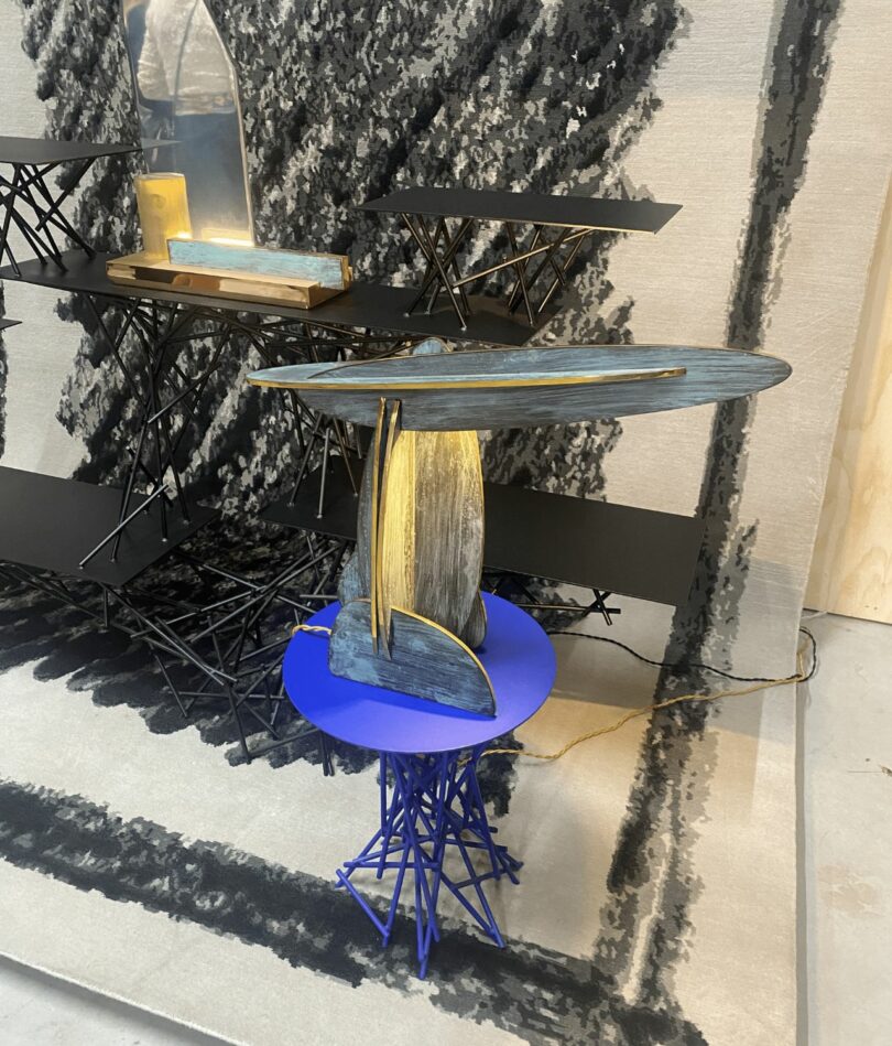 A collection on stools with 'scribbled' style bases made from connecting pieces of metal stand on a rug which is hung on the wall and extends onto the floor. The rug with white with a black scribbled design. On each table is a light made from intersecting flat ovals, edged with gold. 