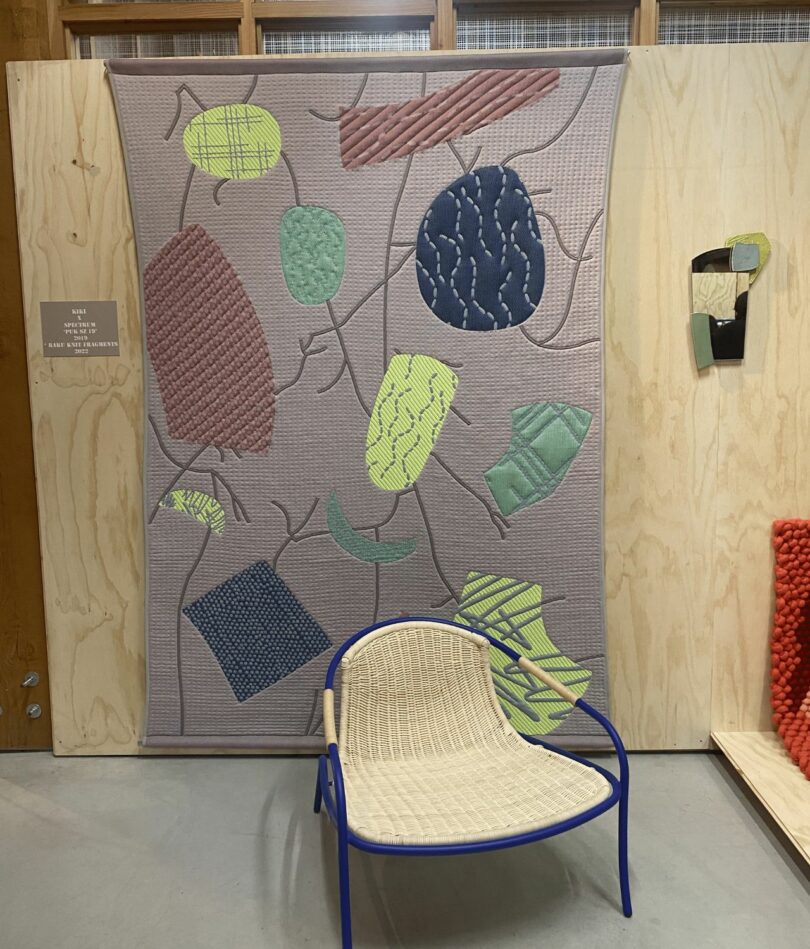 A wall hanging in a lavender colour featured a design made from large irregular shapes in yellow, blue, mint green and a rusty-pink as well as crack-like lines. A low-slung chair is on the floor in front of the wall hanging. 
