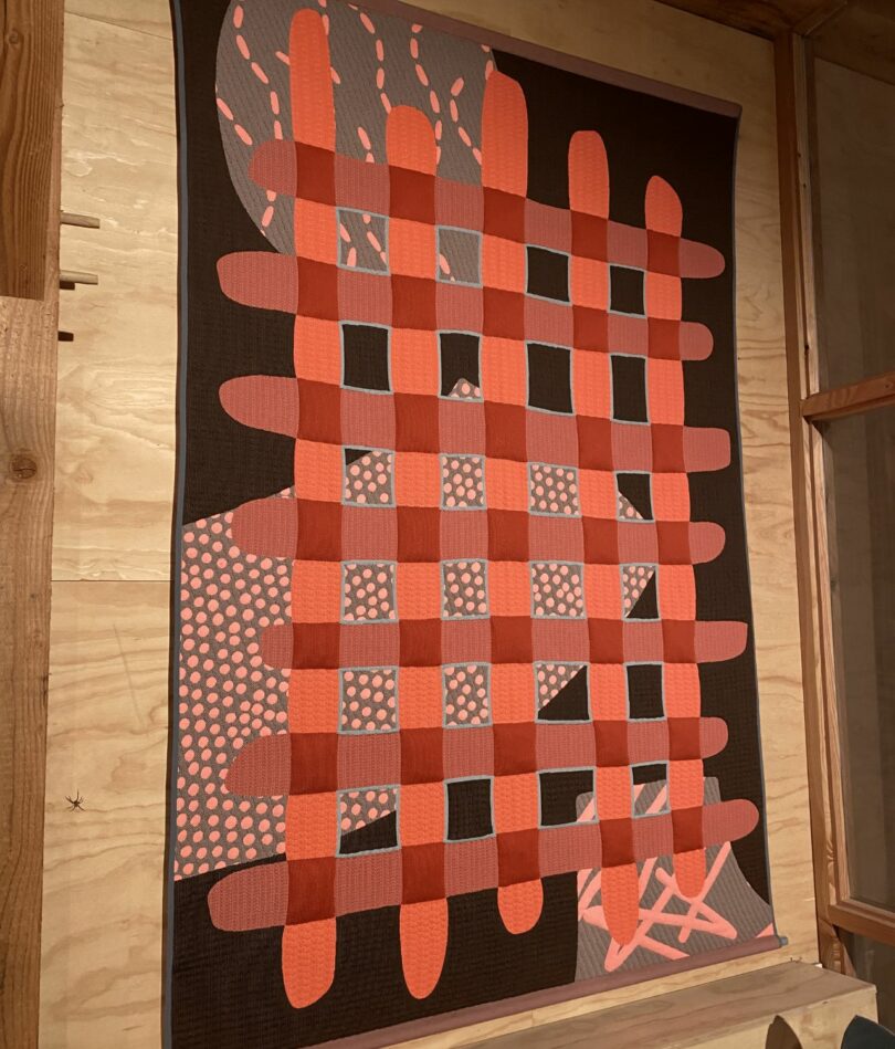 A black wall-hanging featured as red criss-cross design over large areas of grey patterned with pink dots and lines. 