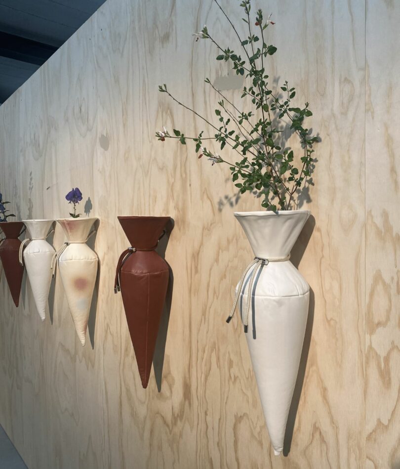 Vases that look like they are made from paper candy bags are affixed to a wooden wall in a row, some have flowers and foliage in them. 