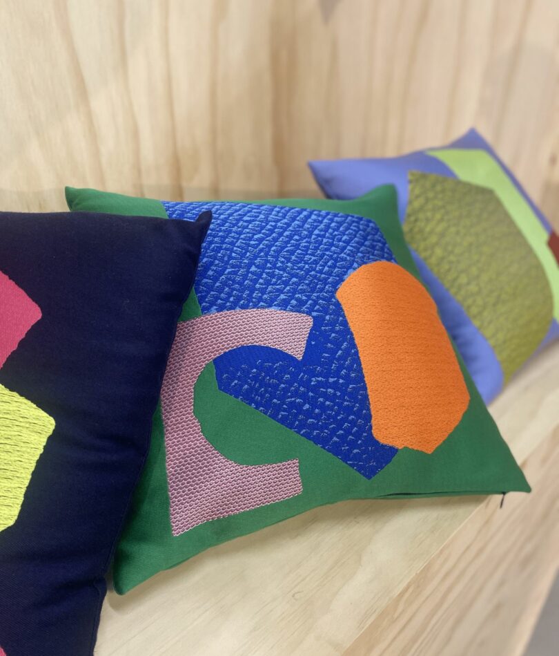 Three scatter pillows are on a wooden shelf. One is black, one is grass green and one is lavender in color. Each has a series of colorful and uneven shapes on the front. 