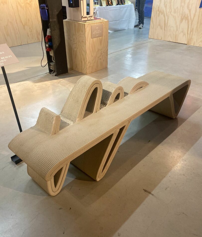 A 3D printed concrete bench features a horizontal surface intersected by a repeating S-shaped line that forms both the legs and the backrest. 