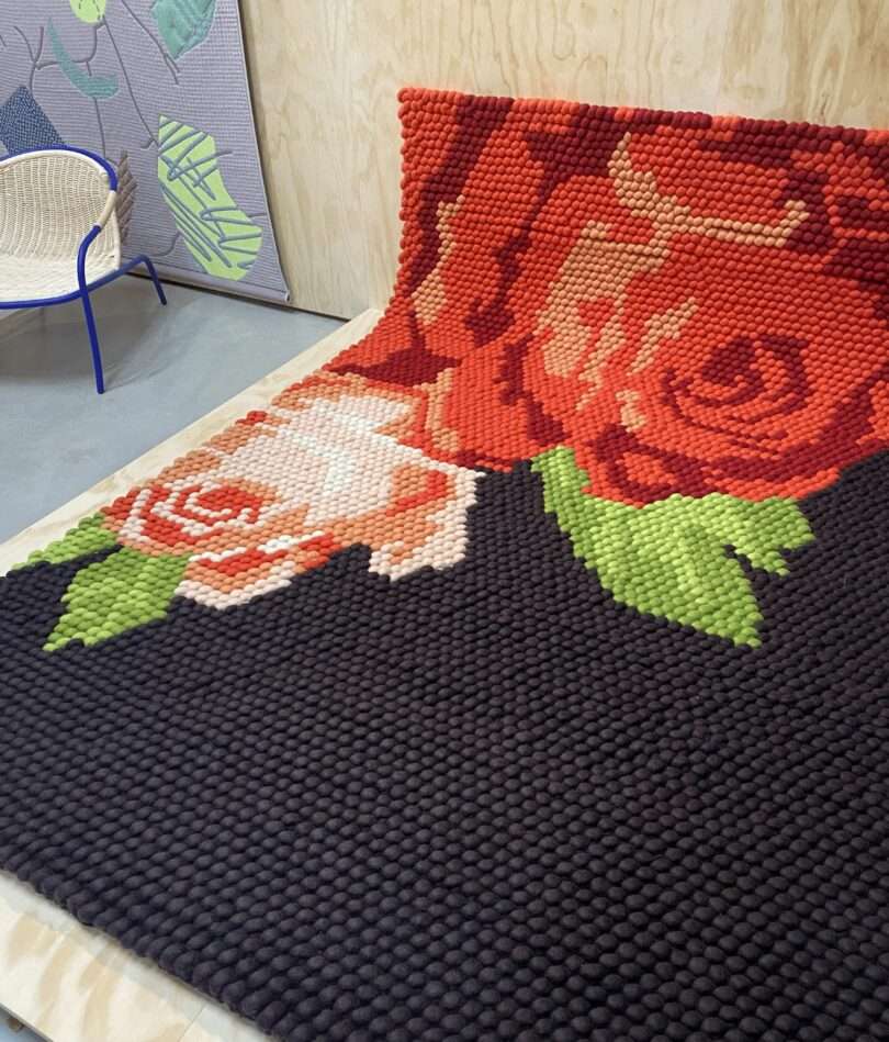 A chunky-knit rug is partially leaning up against a wall. It is black with a large red rose motif. 