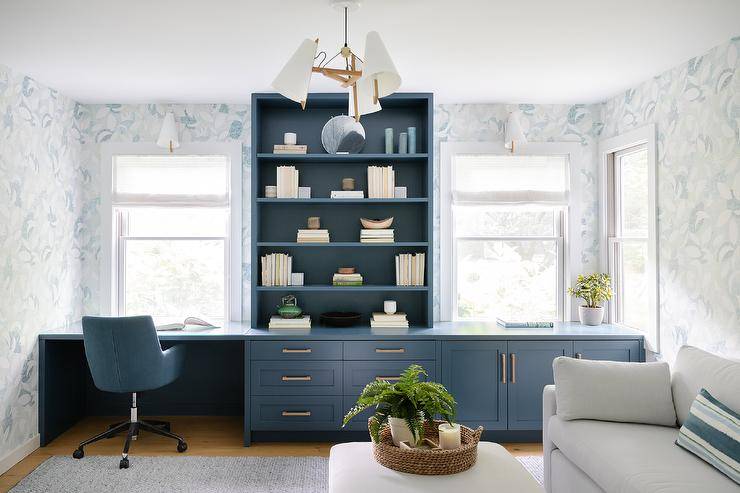 Windows dressed in white roman shades framed by gray and blue wallpaper flank a blue bookcase fixed atop a long blue desk. The desk is finished with blue cabinets donning brass hardware and paired with a blue leather task chair placed on a blue rug.