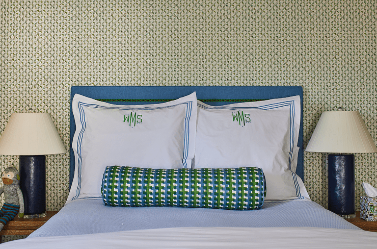 Green and blue monogrammed shams sit in front of a blue nailhead headboard and behind a blue and green bolster pillow placed on blue bedding. Blue lamps sit atop brown wooden nightstands.