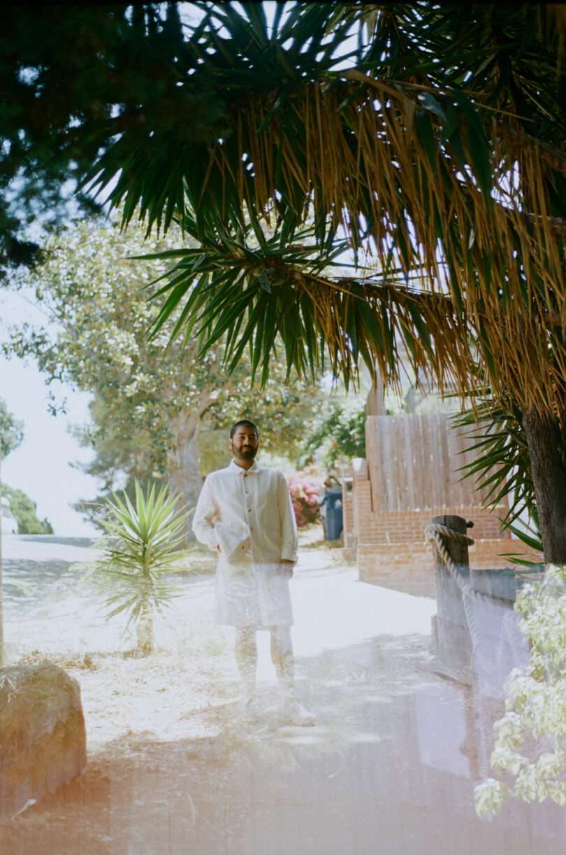 brown-skinned man dress in all white stands under a large tropical tree