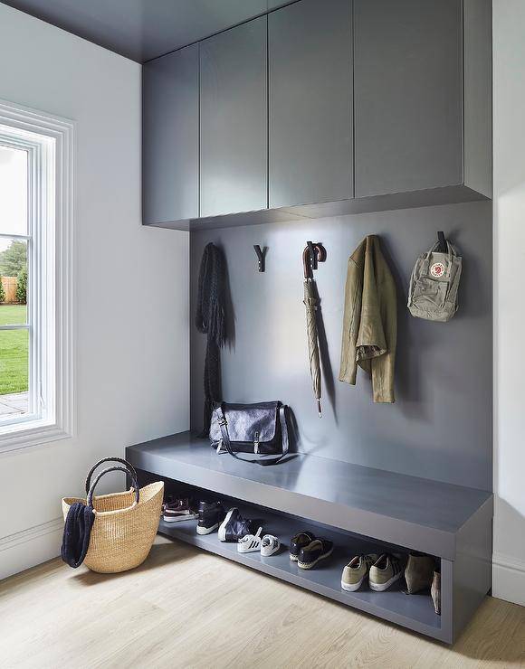 A dark gray mudroom bench with a built-in shoe shelf is fixed against a gray backsplash and under gray flat front cabinets.