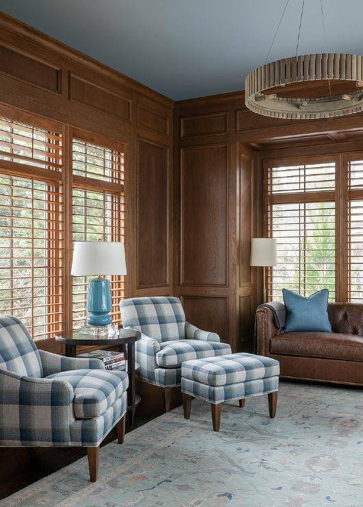Welcoming oak paneled home office features blue plaid chairs placed on either side of a round brown accent table topped with a blue lamp, while one chair is paired with blue plaid bench positioned on a blue vintage rug. A wooden ring chandelier hangs from a blue painted ceiling.