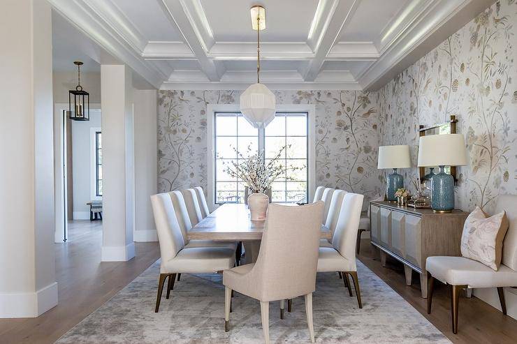 Aerin Culloden Lamps atop a brown prism buffet cabinet adjacent to a gray wash wooden dining table fitted with mixed chairs in taupe and white. This transitional dining room boasting coffered ceilings and floral wallpaper for a charming traditional finish.
