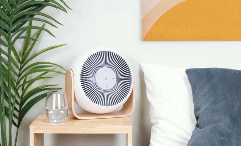 Snooz Breez Fan and Sound Machine with bent plywood base stand set on top of a light wood bedside table with a glass of water, set near a bed topped with white and blue pillows, yellow art on the wall, and a faux palm plant to the right.