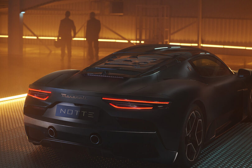 Rear view of Maserati MC20 Notte Edition super sports car in matte and glossy black finish with "NOTTE" license plate and two figures in the background bathed in hazy orange light.