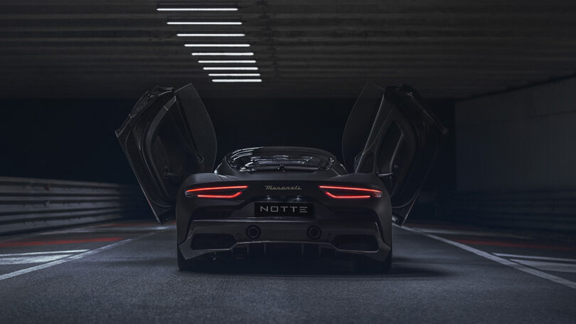 Rear view of Maserati MC20 Notte Edition super sports car in matte and glossy black finish with "NOTTE" license plate and both gull wing doors angled open, parked inside darkened underground parking.