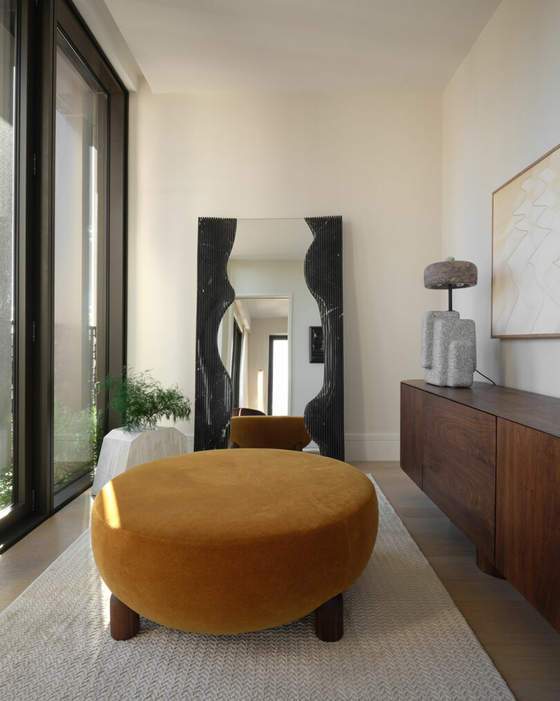 A mustard colored ottoman in the foreground of a minimal designed interior.