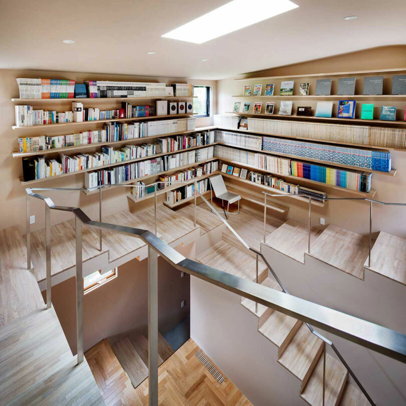 angled down interior view of boxy modern house with book shelves lining open perimeter and stairs wrapping around the sides.k