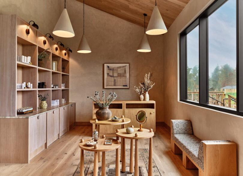 Organic decorated interior with large wood floor to ceiling shelves, small narrow ottoman daybed and a trio of nesting circular tables in the center. Four cone pendant lamps overhead.