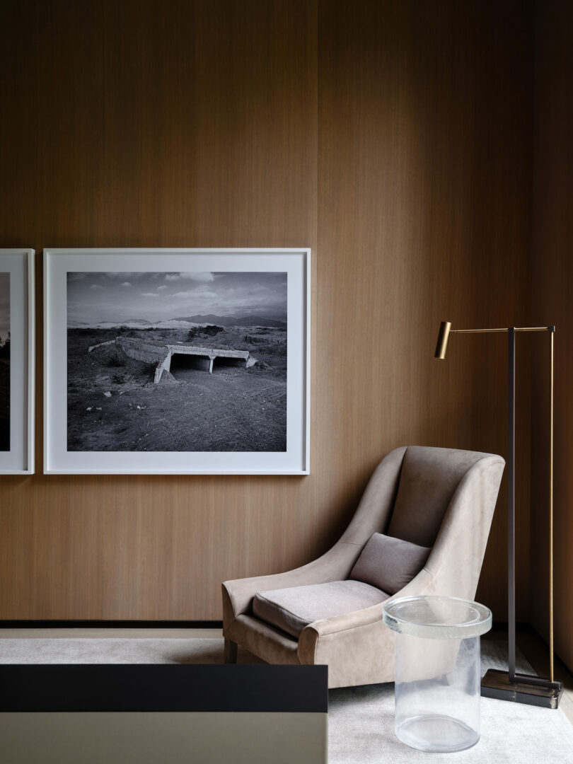Wood walls with two large black and white photographic framed prints behind upholstered arm chair and clean side cylindrical side table with minimalist floor lamp behind the chair.