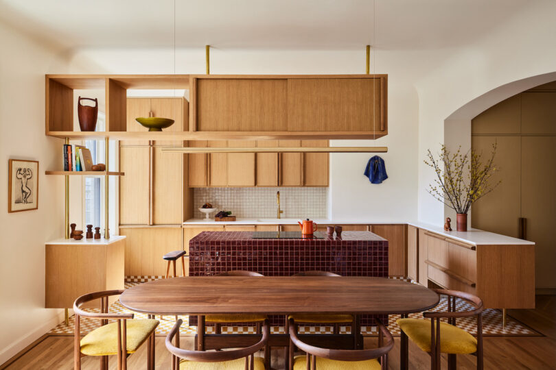 Warm modern kitchen with dark burgundy tiled kitchen island and wood overhead shelving. Dining table with 6 midcentury seats in the foreground.