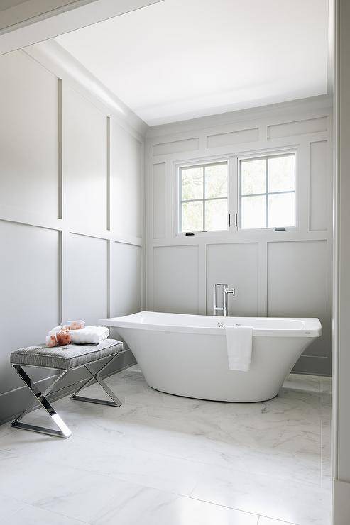 Floor to ceiling light gray wainscoting in master bathroom adds a classic finish to the walls completed with a modern freestanding tub paired with a floor mount chrome tub filler. A gray X bench furnishes the bathroom against the wainscoting wall and bathtub styled to complement the chrome and gray finishings.