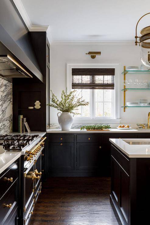 Stylish black kitchen boasts black cabinets donning brass knobs and holding a sink with a brass gooseneck faucet beneath glass and brass shelves.