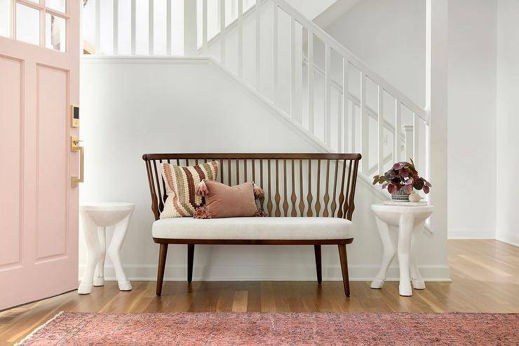 A pink front door opens to a foyer boasting a pink runner placed in front of a vintage wooden bench flanked by white hoofed accent tables placed against a staircase wall.