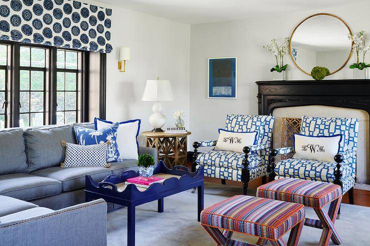 Mixed patterns, color schemes, and design furnishings come together beautifully to create a contemporary living space. A blue denim sectional with nailhead trim is decked with a few blue chinoiserie pillows next to a blue Charleston Coffee Table on a light blue area rug. White and blue geometric print chairs mismatch next to colorful striped x stools but manage to contrasts one another in a bold yet fitting combo. Black wooden fireplace mantle with a brass arabesque screen gives a platform for a lovely large gold mirror flanked by two floral arrangements in a white square vase. A brass and cream Aspect Sconce is mounted next to a black framed window dressed in blue and white pattern shade over a round wood end table featuring a white double gourd lamp.