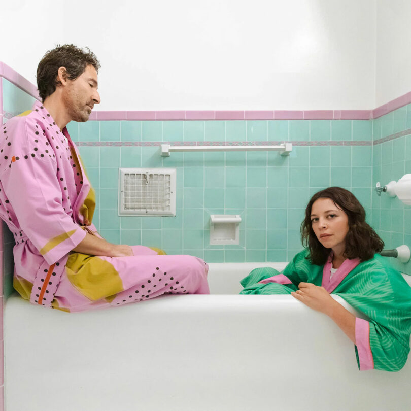 Two people sitting in and on a bathtub while wearing robes.