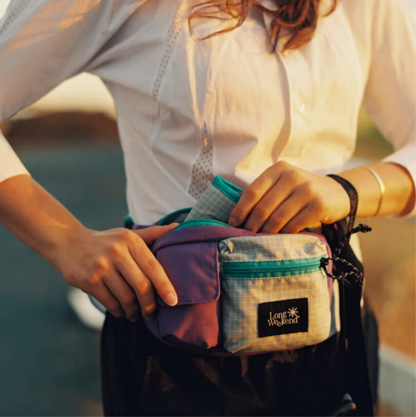 Woman in white button up long sleeve shirt pulling out a Padded Camera Wrap from her Long Weekend sling with warm sunset light across her torso.