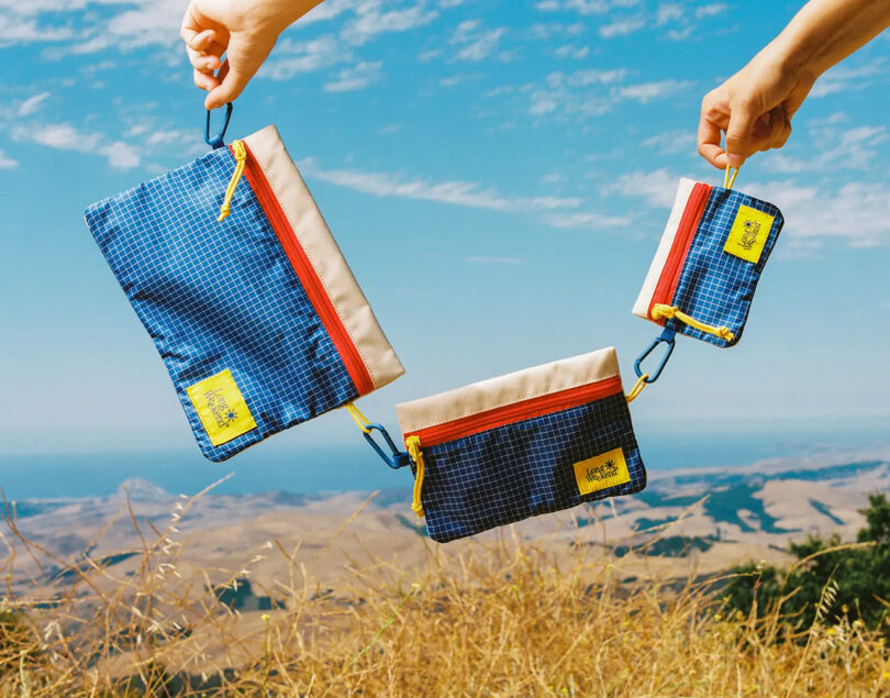 Small, medium and large Long Weekend Everyday Zip Pouches interlinked by carabiner clip being and held up outside with rolling hills and blue skies in the background.